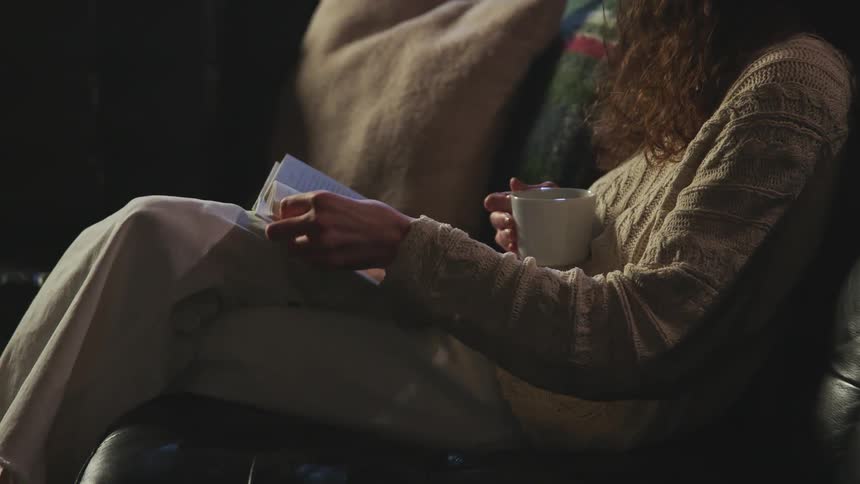 Woman comfortably sitting on a couch at home reading a book with a cup on her hand
