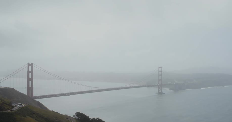 Automobiles travel on a road leading to a suspension bridge stretching across the water and into the fog on the other side