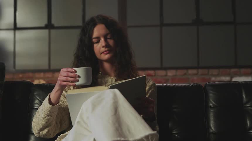A woman reaches for her mug of coffee and drinks while reading a book on the couch