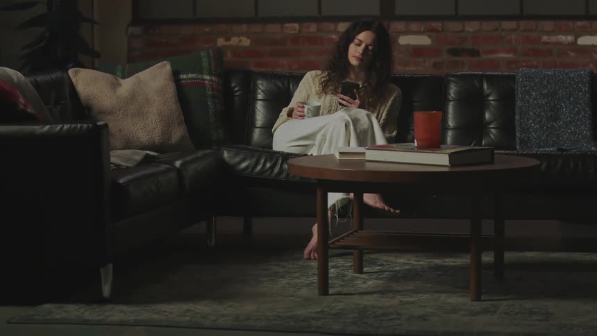 Woman with curly hair beige shirt and white pants sits on black couch using smartphone in living room