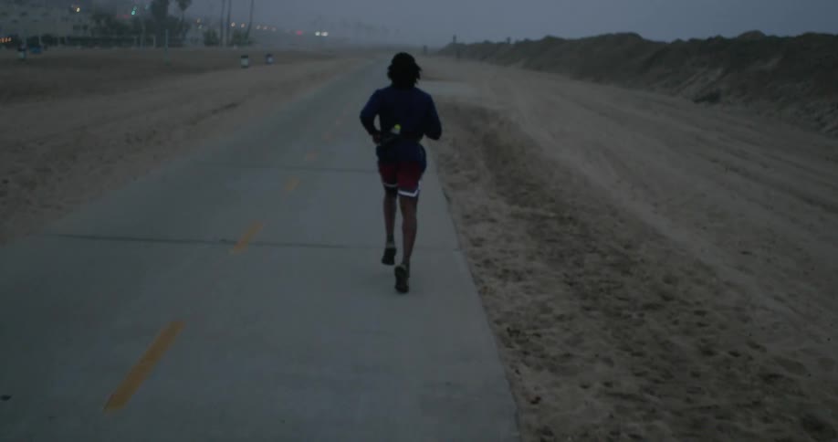 Man with dreadlocks black jacket red shorts and running shoes jogs on coast road