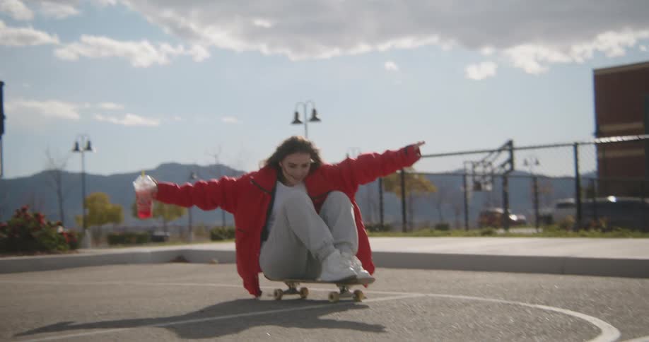 A girl is holding a drink and sitting on a skateboard as it rolls across a playground