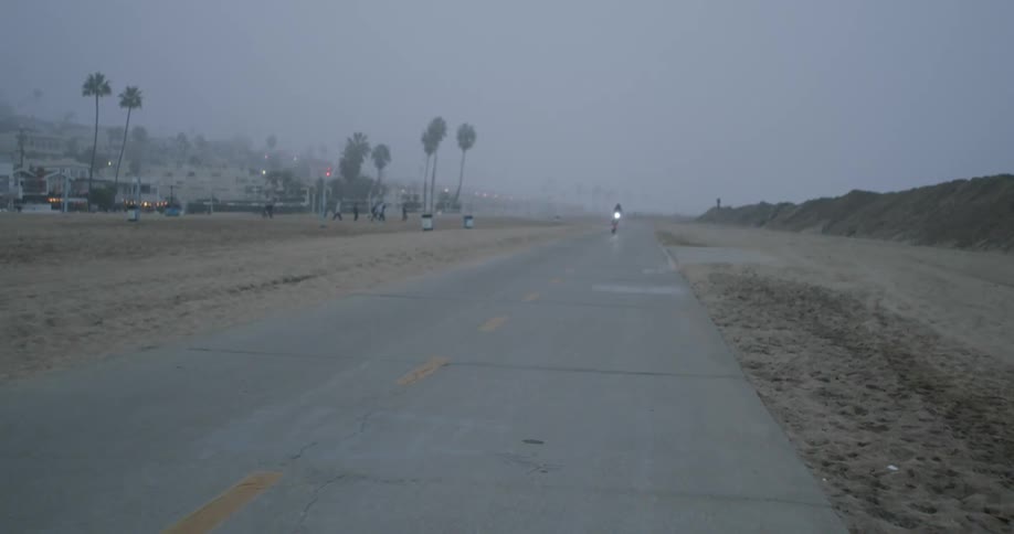 A person drives an electric scooter on a road in a beach near a small town under the mist