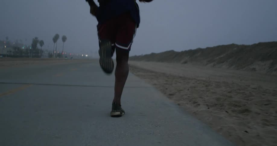 Man with dreadlocks black jacket red shorts and running shoes jogs on coast road