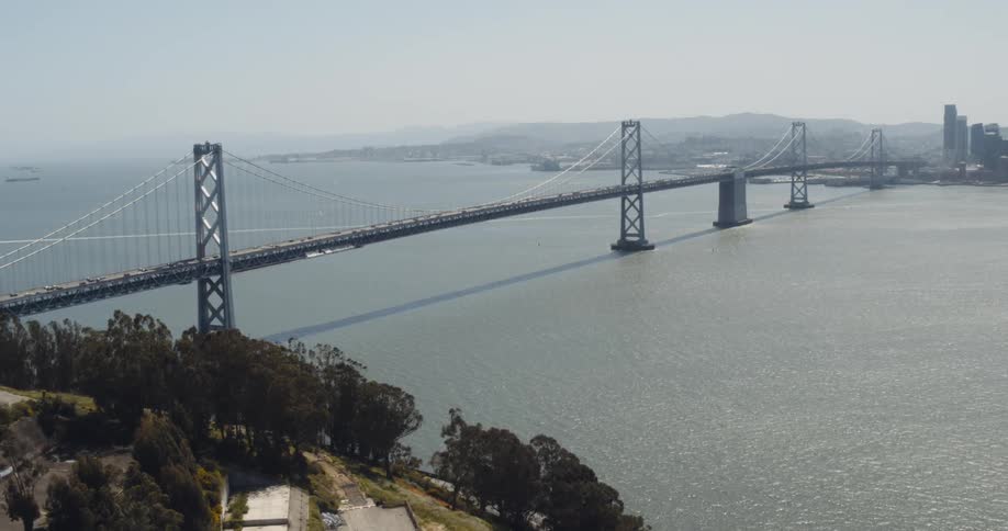 The bay bridge with skyscrapers at one end and hills and trees at the other