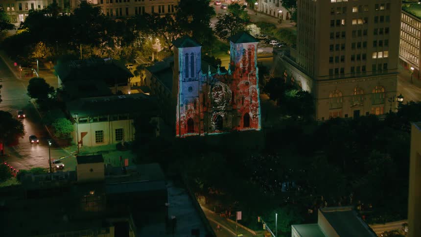 Traffic flows along city streets at night passing event with people in parking lot viewing image projected onto building