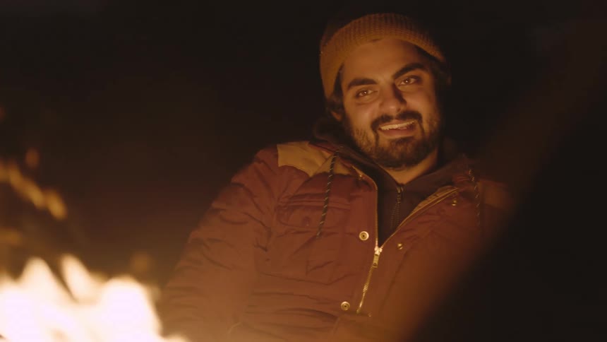 Young man with beard sits next to campfire smiling