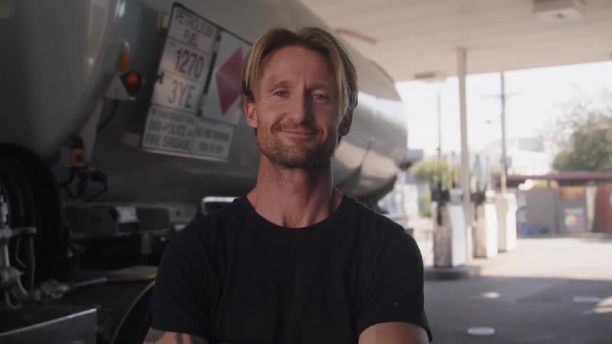 A man is standing outside at a gas station near a truck and he smiles