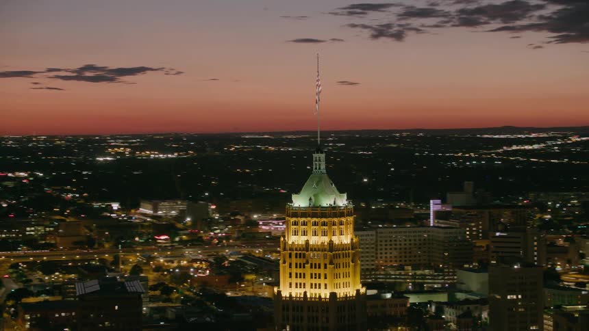 An american flag flies on top of a building that is lit up as the sun sets overlooking a big city