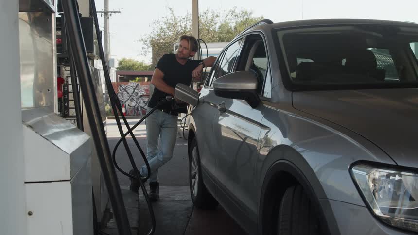 A man is standing next to an automobile as he fills it with gas and puts the gas hose back on the pump