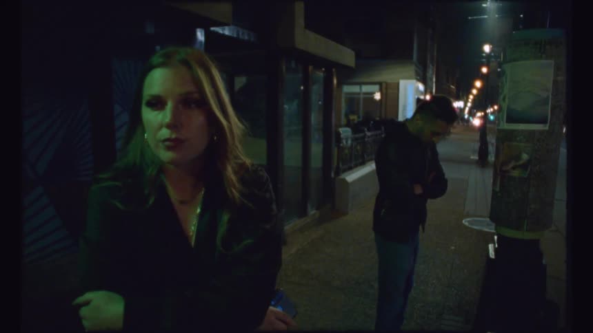Woman holding smartphone and man stand waiting near smoking man leaning against building in city at night