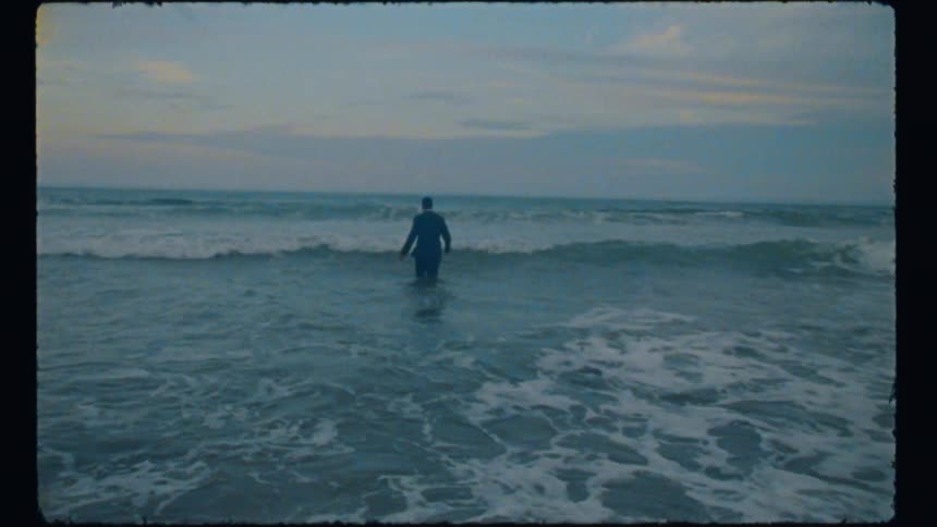 A man wearing a business suit walking through the waves into the ocean