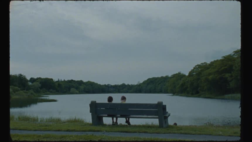 Two children sit on bench overlooking lake across from woods and near path