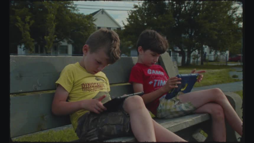 Two young boys are sitting on a neighborhood bench playing with electronic tablets