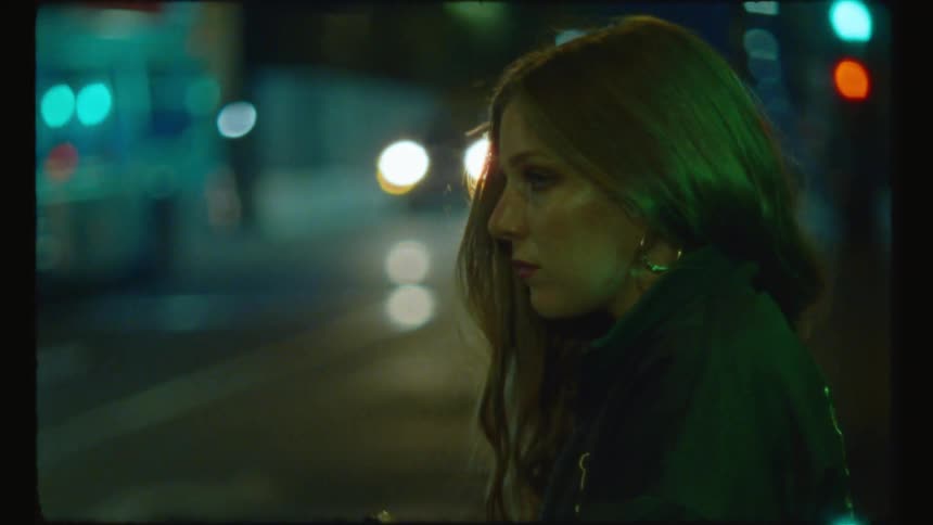 Woman stands near another as they both wait near busy city street at night
