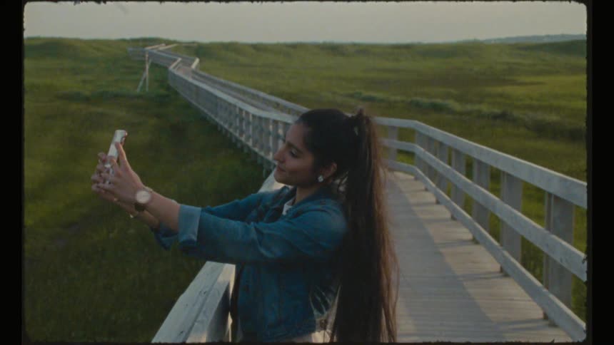 A teenage girl stands on a bridge in a grassy field and takes a selfie with her phone
