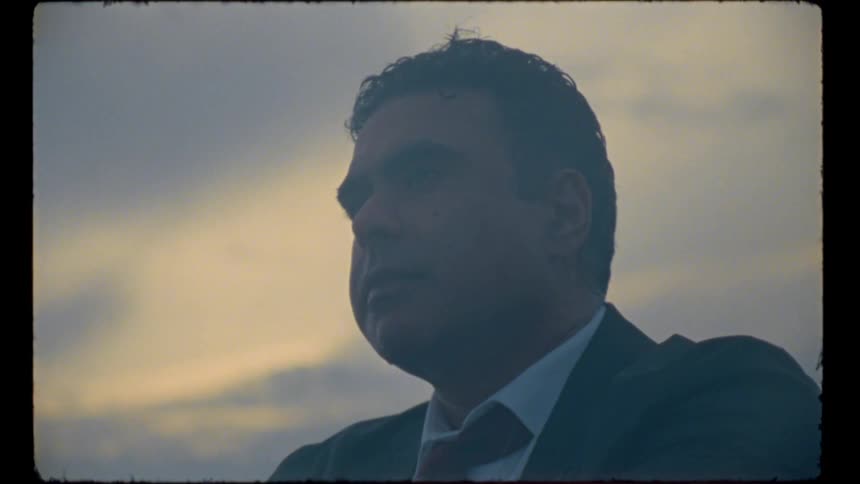 A man in a suit sits beneath a cloudy sky and pensively looks off into the distance