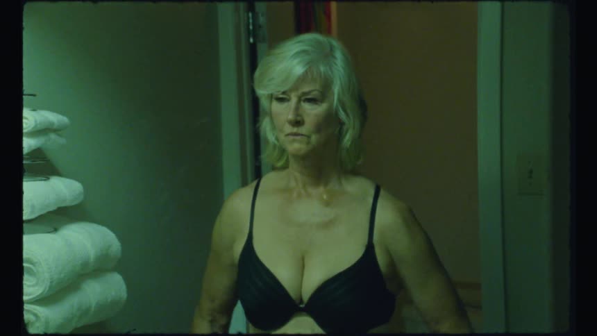 An elderly woman stands in front of a mirror wearing nothing but her underwear while looking at her own reflection