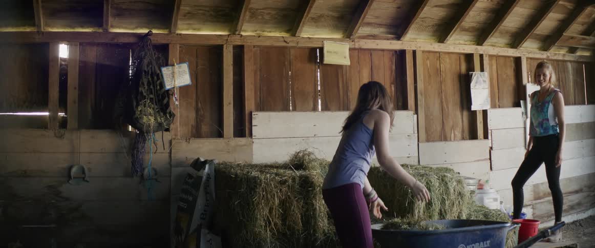 A woman stands in a barn and watches while another woman places hay into a wheelbarrow