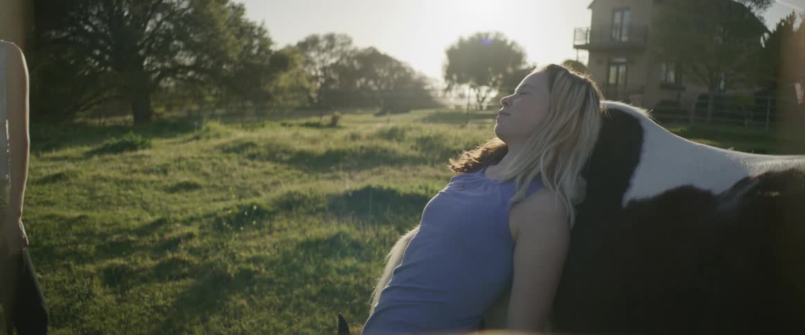 A teenager lies against her horse while her mom watches nearby