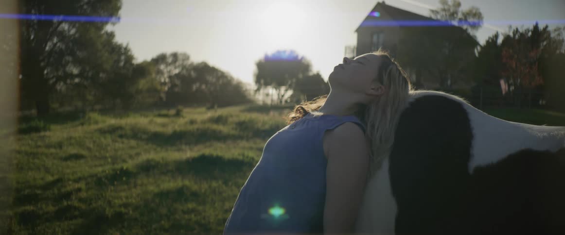 Woman leans against side of horse on sunny day with house in the background