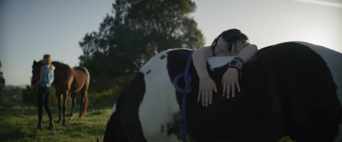 Girl leans over horses back patting the animal as another woman stands next to a second horse in field on sunny day