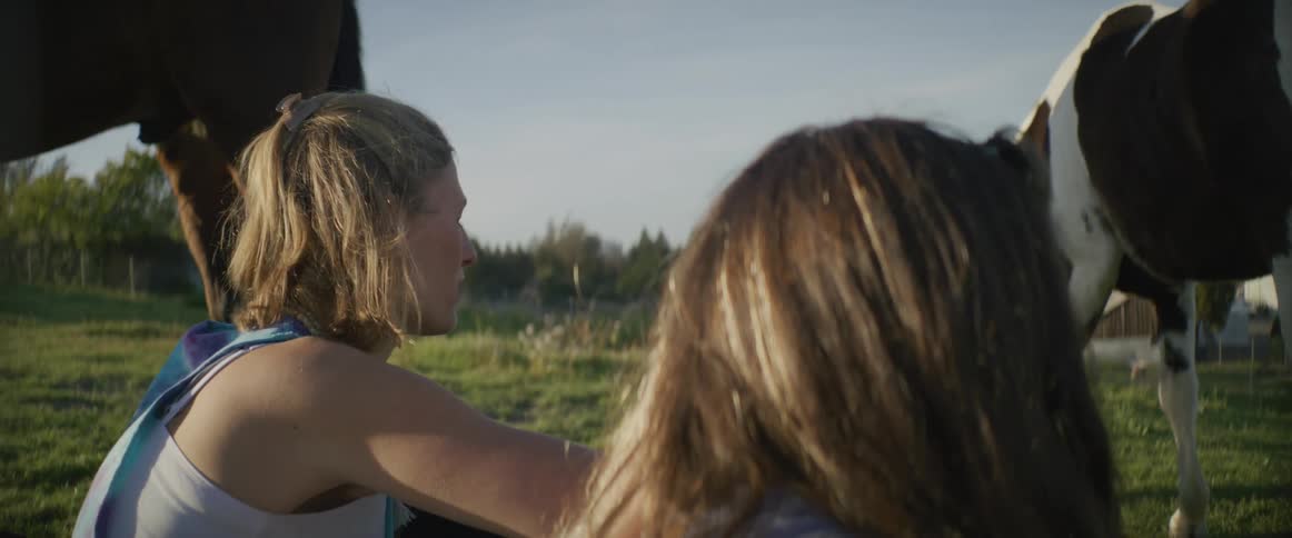 A mother and daughter sit in a grassy field with two horses as they graze