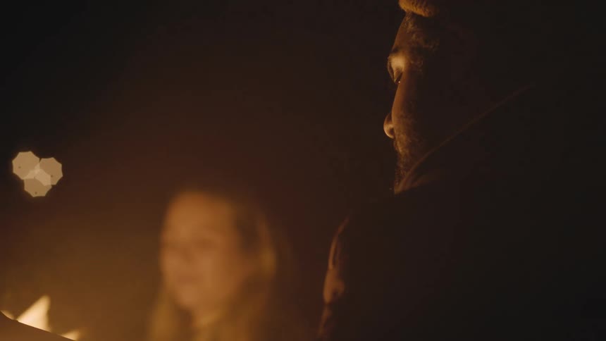 A woman sits by a fire and friend who sings and plays the guitar