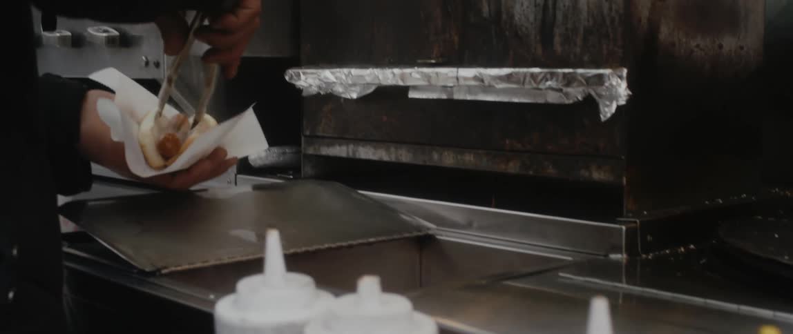 A man in a restaurant puts a hot dog in a bun for a customer