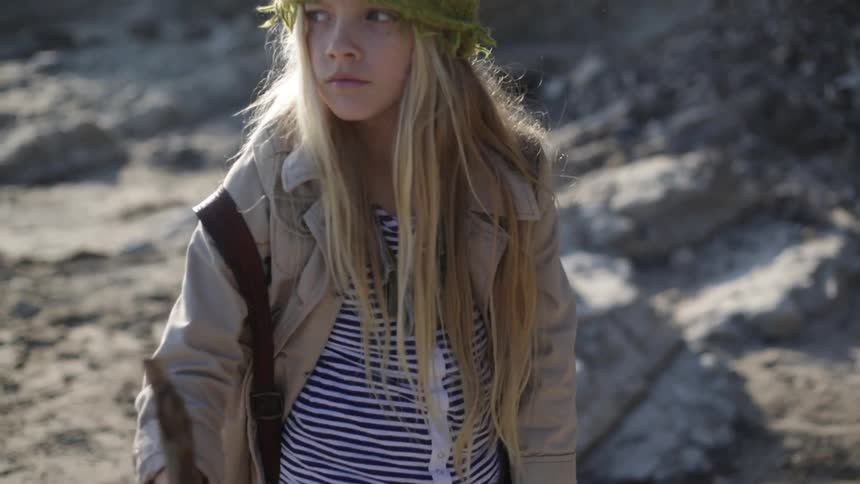 A girl is walking along some rocks and sand holding a stick