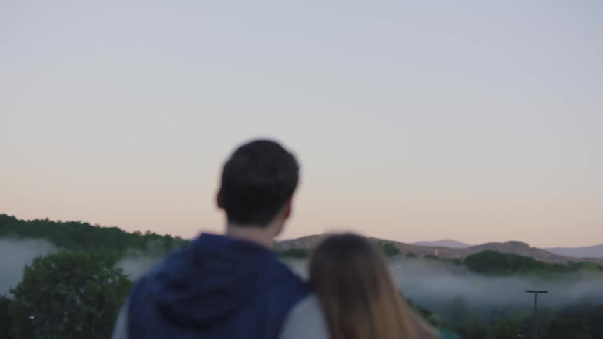A woman standing with her head leaning on her boyfriends shoulder as they look out at the fog above the trees
