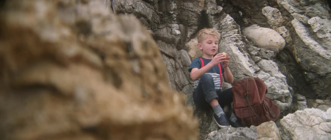 A boy is sitting on some rocks pulls an apple out of his bag and eats