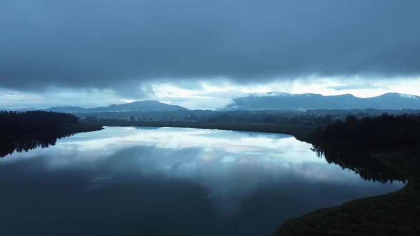 A cloud filled sky reflects on a lake surrounded by forests with mountains in the distance