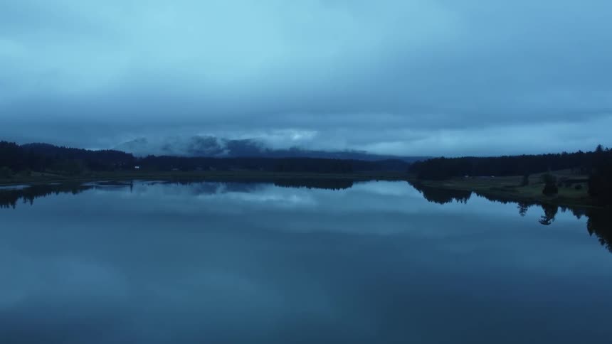 Mirror like surface of lake reflects nearby forest overcast sky and mountains