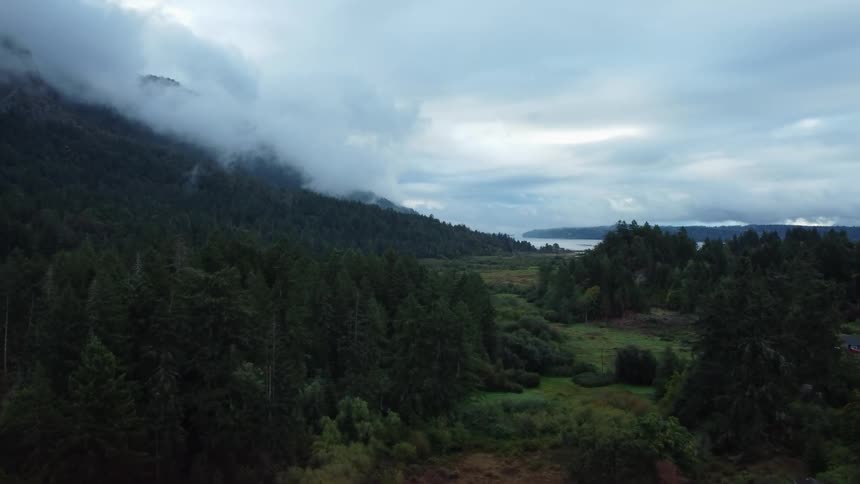 Vehicles drives past house near forest covered mountain with body of water in background