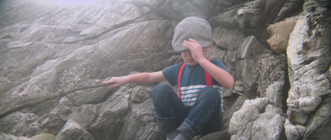 A boy sits down on rocks holding a branch