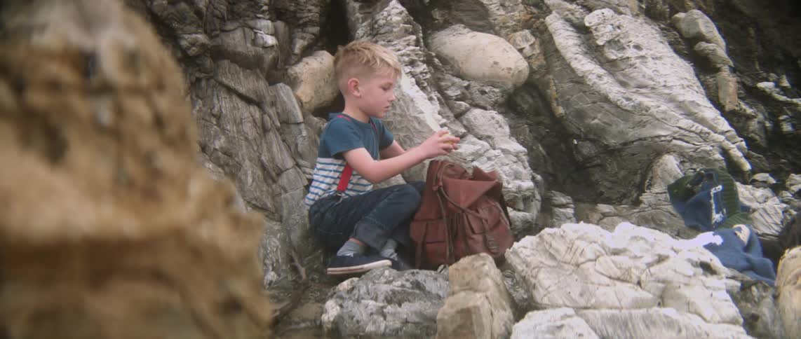 A boy sitting on some rocks opens his backpack to get something to eat