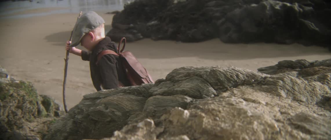 A boy carrying a stick is climbing on rocks by the beach
