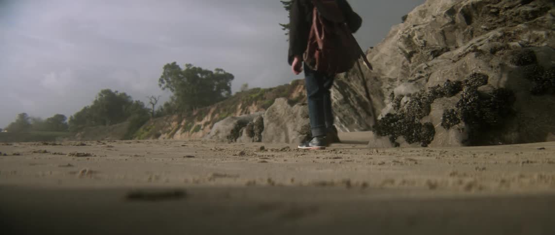 A boy walks by some rocks with a stick in hand