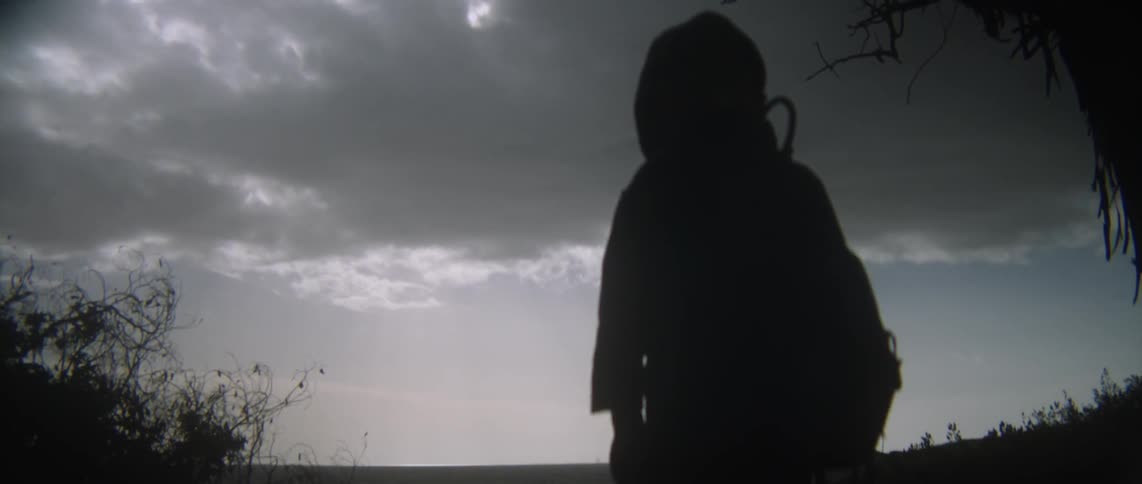 A child with a stick is standing by the water looking up at dark clouds above