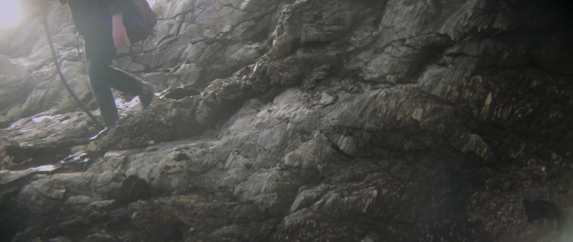 Person walks over rocks on the beach holding a walking stick