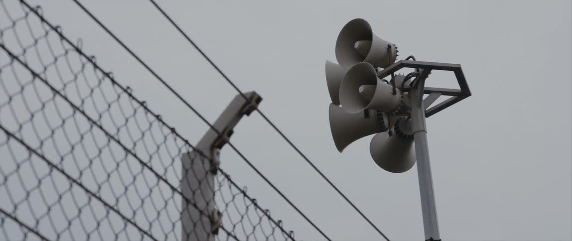 Several loudspeakers on a single post inside a tall security fence