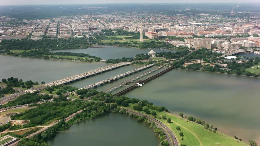 A jet flies over traffic covered bridges preparing to land with the washington monument and city behind it
