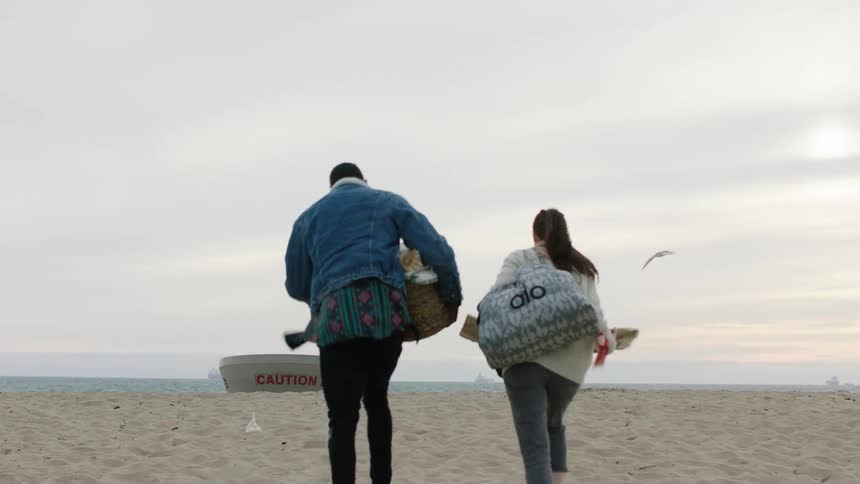 A man and a woman are walking away from a car parked on a beach and they are carrying bags