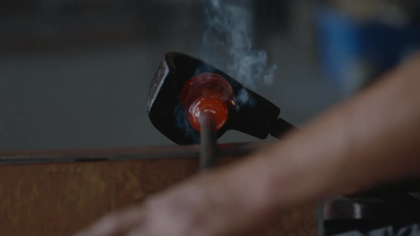 A glassmaker is rolling molten glass on the end of a pole