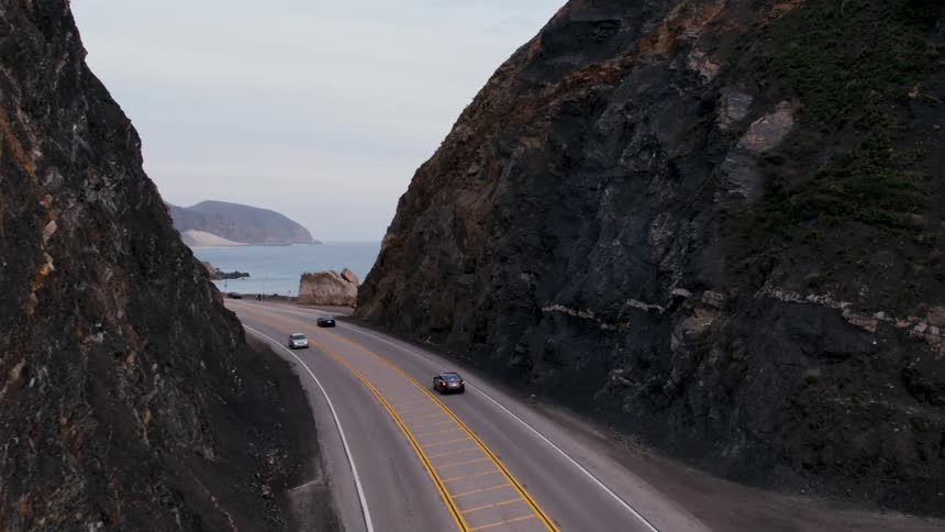 Automobiles travel on a road between mountains along the coast