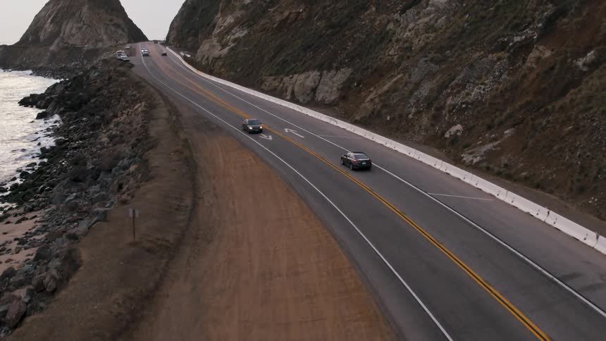 Automobiles travel on a road along the coast that wraps around a mountain as waves roll over rocks below