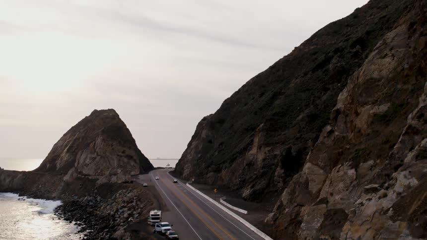 Automobiles travel on a road along the coast that wraps around a mountain as waves roll over the beach and the sun shines on the water