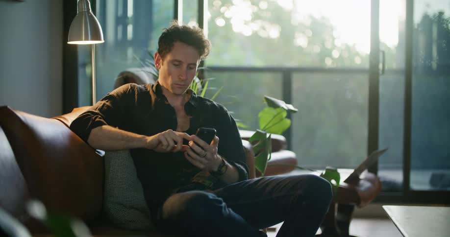 A man uses a smartphone while sitting on a sofa indoors during the day