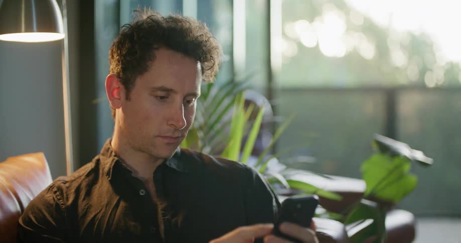 A man is sitting on a couch in a living room using his cell phone